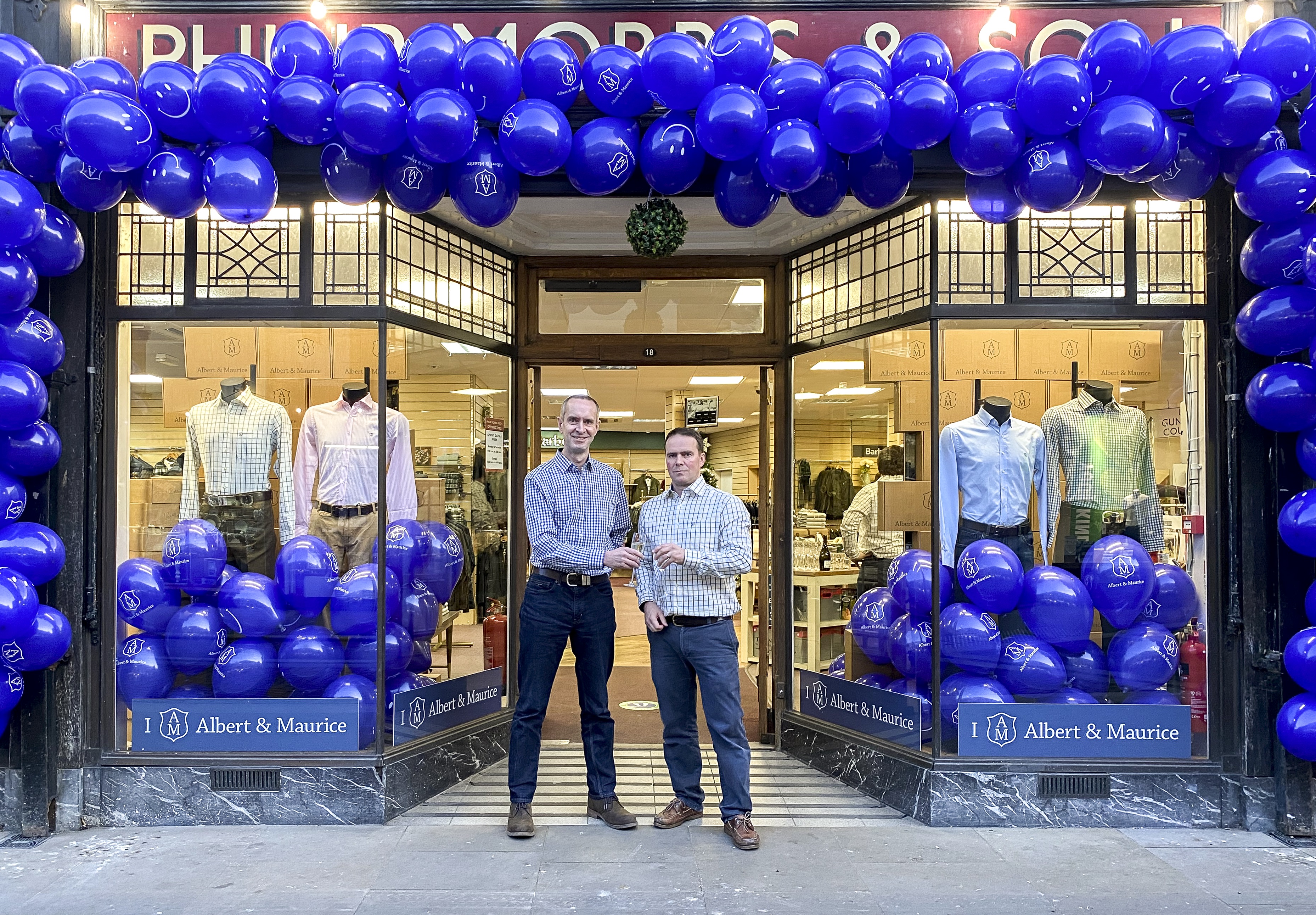 John and Bruce celebrate the launch with a glass of bubbly. Blue balloons with the A&M logo frame the outside of the shop. 